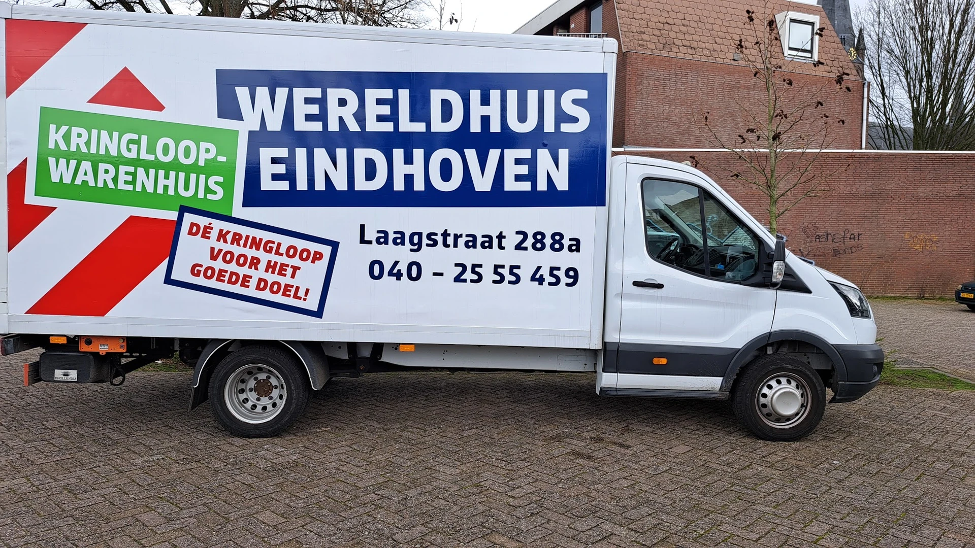 Witte bedrijfsbus met blauwe opdruk 'Wereldhuis Eindhoven' en contactgegevens op straat geparkeerd