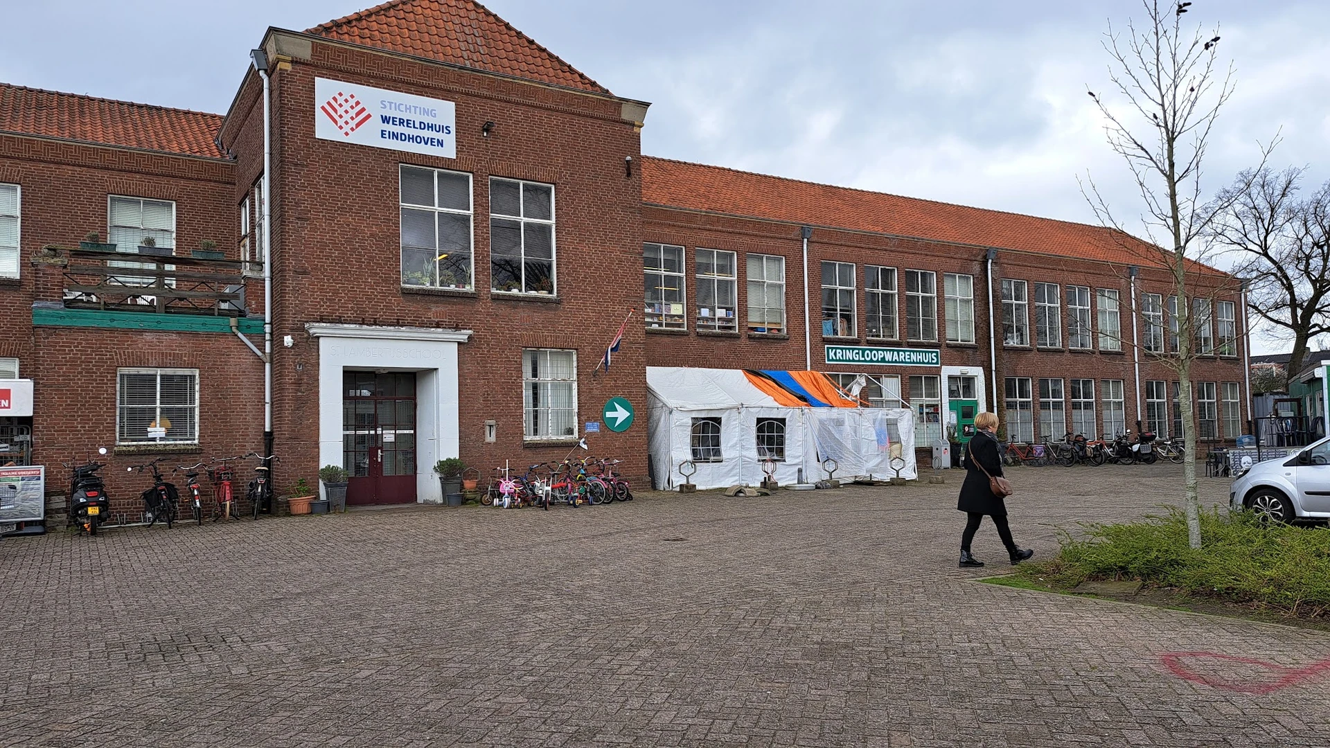 Voorkant van bakstenen kringloopwinkel met winkelnaam bord, rode dakpannen en wit dekzeil over deel van gevel