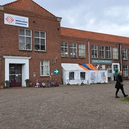 Voorkant van bakstenen kringloopwinkel met winkelnaam bord, rode dakpannen en wit dekzeil over deel van gevel
