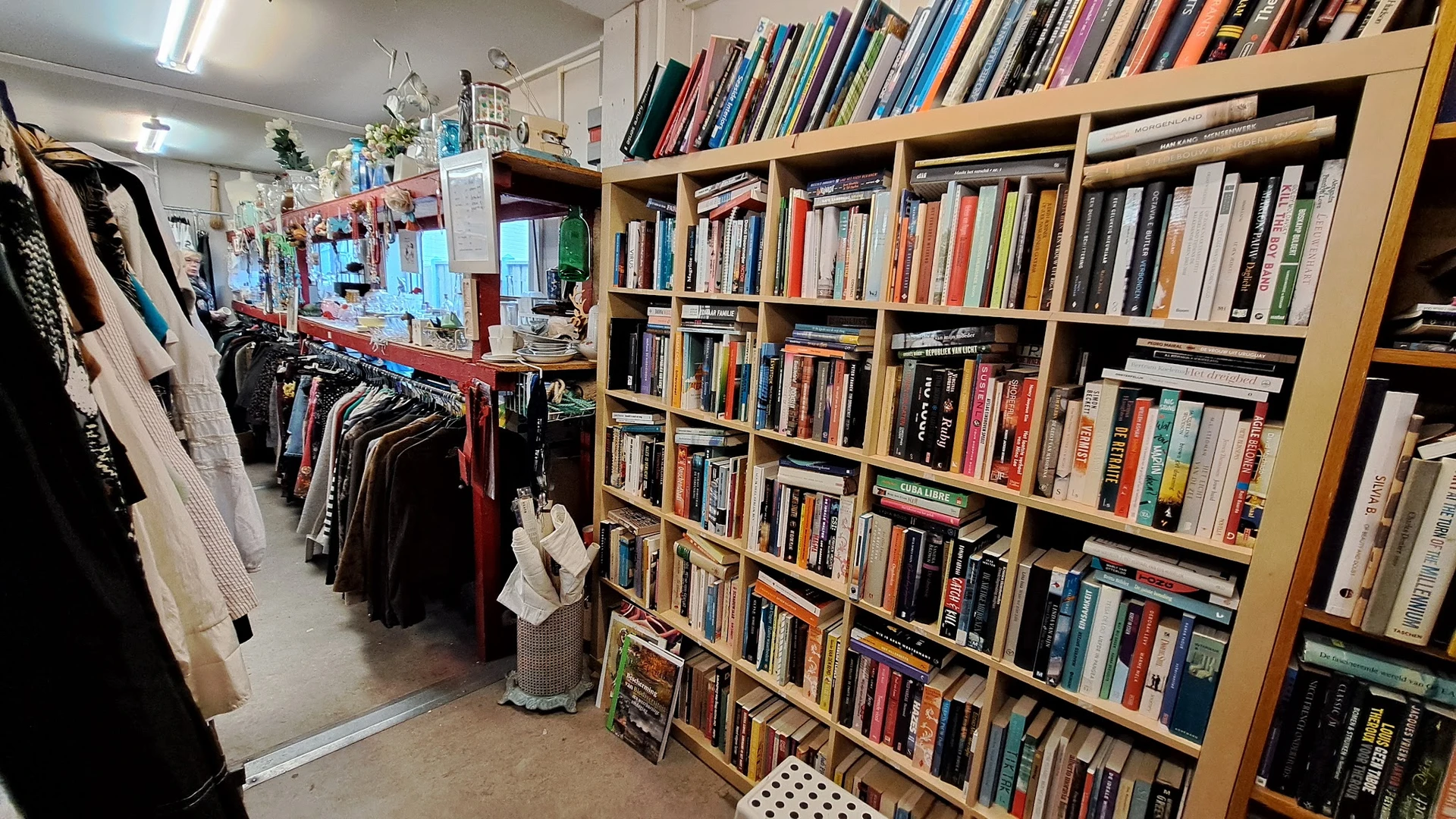 Grote boekenkast met honderden tweedehands boeken in verschillende kleuren, links ervan kleding op hangers