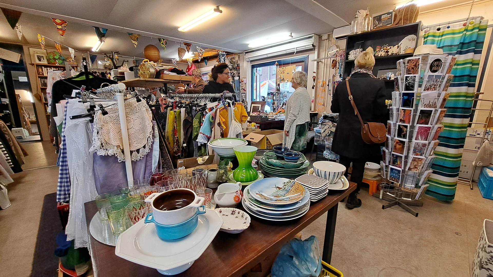 Winkelbinnenkant met tafels vol servies, kleding aan rekken, klanten aan het winkelen, volle schappen tegen muren