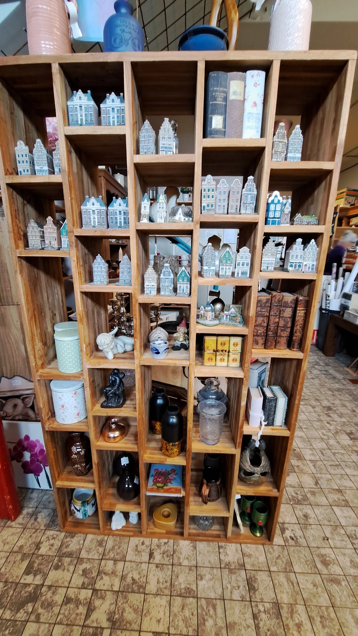 Houten wandkast met vakken vol decoratieve items: miniatuurschilderijtjes van huizen, porseleinen beeldjes, zwarte kettels, k