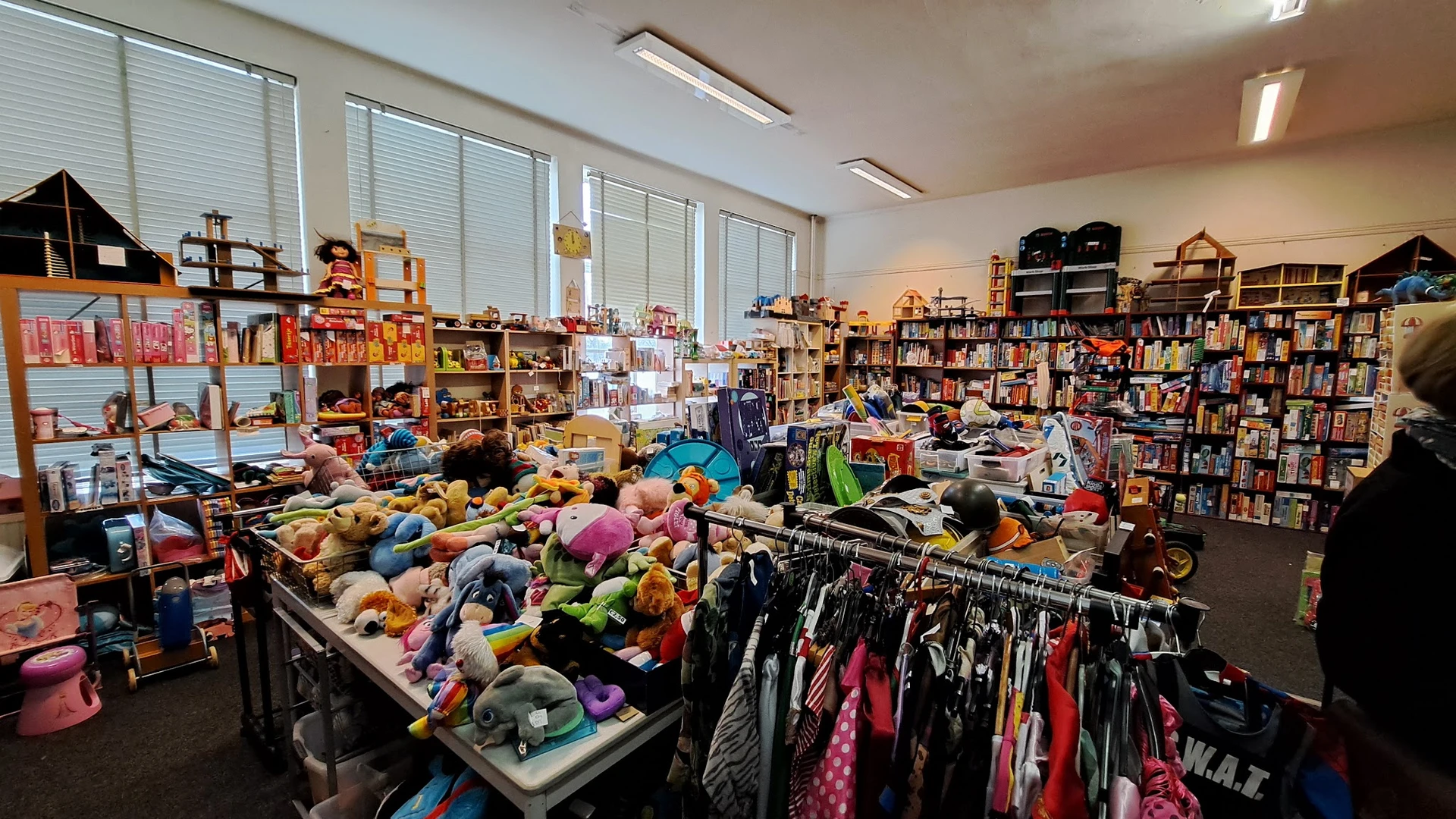 Winkelinterieur met speelgoed, kleding en boeken; tafels vol knuffels en pluche dieren, rek met kinderkleding, boekenkasten o