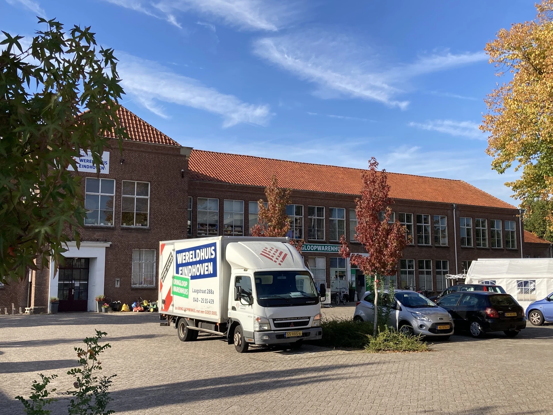 Bakstenen gevel van kringloopwinkel met oranje dakpannen, parkeerplaats met witte bedrijfsbus en auto's ervoor
