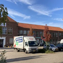 Bakstenen gevel van kringloopwinkel met oranje dakpannen, parkeerplaats met witte bedrijfsbus en auto's ervoor