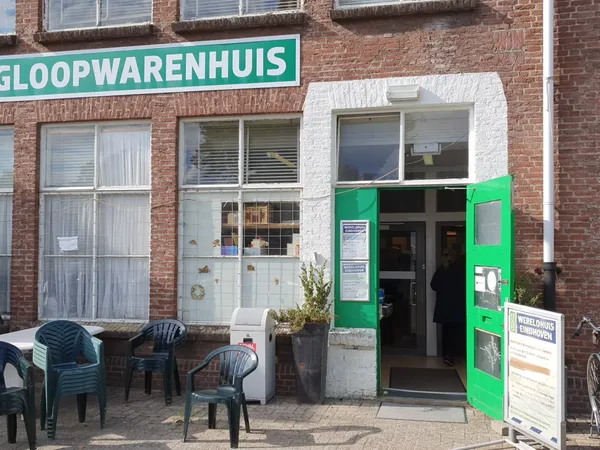 Voorgevel van kringloopwinkel met groene deur, wit kozijn en turquoise winkelnaambord, terras met stoelen ervoor