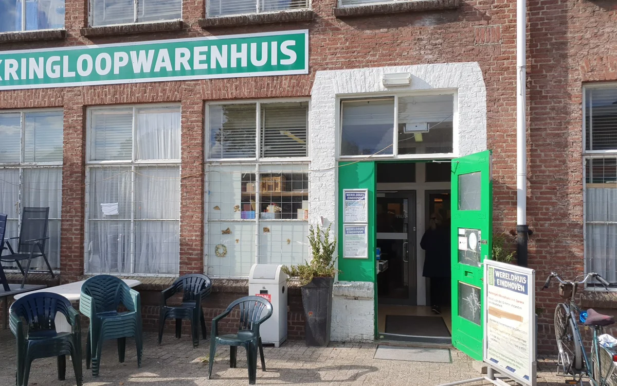 Voorgevel van kringloopwinkel met groene deur, wit kozijn en turquoise winkelnaambord, terras met stoelen ervoor