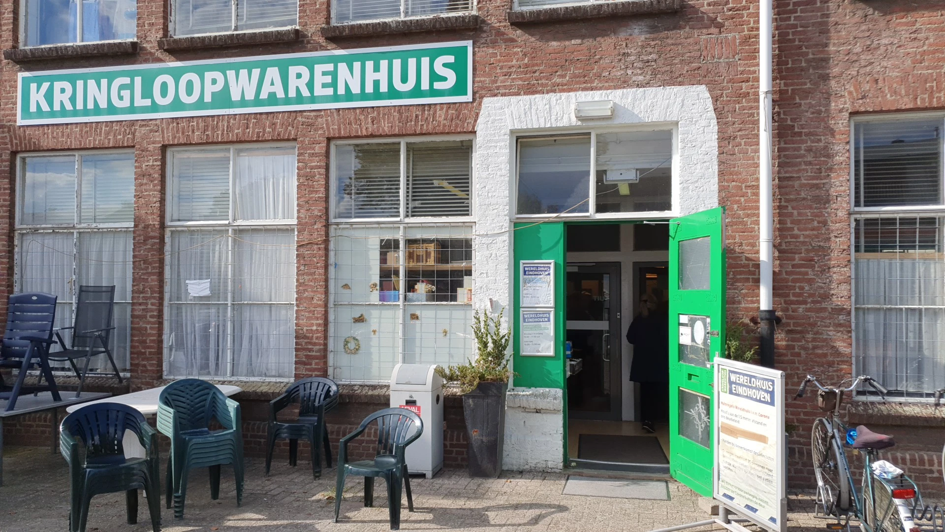 Voorgevel van kringloopwinkel met groene deur, wit kozijn en turquoise winkelnaambord, terras met stoelen ervoor