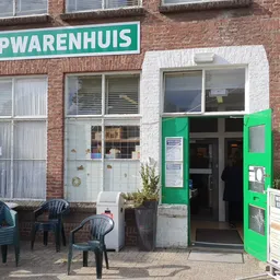 Voorgevel van kringloopwinkel met groene deur, wit kozijn en turquoise winkelnaambord, terras met stoelen ervoor