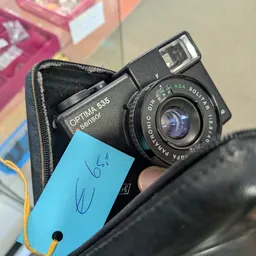 Close-up van een vintage fotocamera Optima 535 met flitsapparaat in een zwarte tas, naast een blauwe label