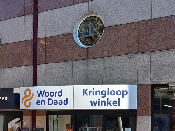 Gevel van kringloopwinkel Woord en Daad met horizontaal winkelnaambord, rood-wit gevelsteen en ronde raampje.