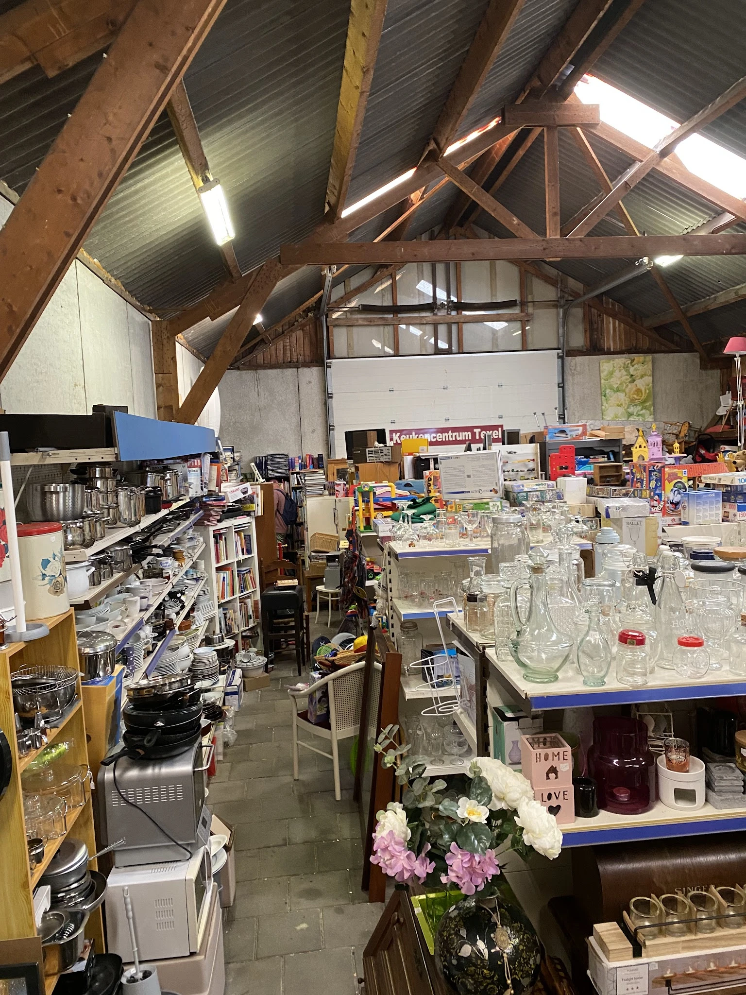Overzicht van een kringloopwinkelruimte met houten dakspanten, stellingen met diverse spullen, glazen potten en huishoudartik