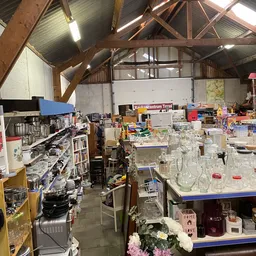 Overzicht van een kringloopwinkelruimte met houten dakspanten, stellingen met diverse spullen, glazen potten en huishoudartik