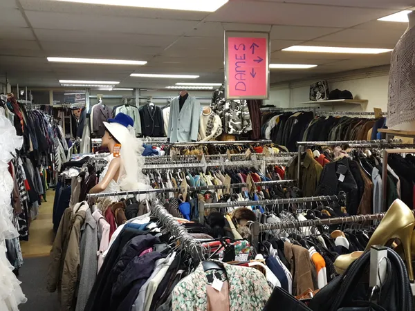 Overzicht van de winkelvloer met meerdere rekken vol tweedehands kleding, een roze uithangbord zichtbaar boven