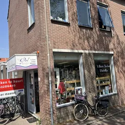 Bakstenen gevel van kringloopwinkel met etalageramen, fiets ervoor, winkelnaam zichtbaar op raam