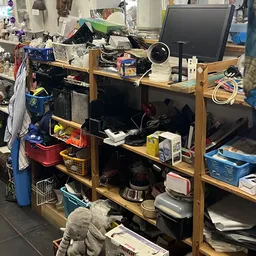 Rommelige winkelruimte met houten stellingen vol elektronische apparatuur, dozen en diverse spullen