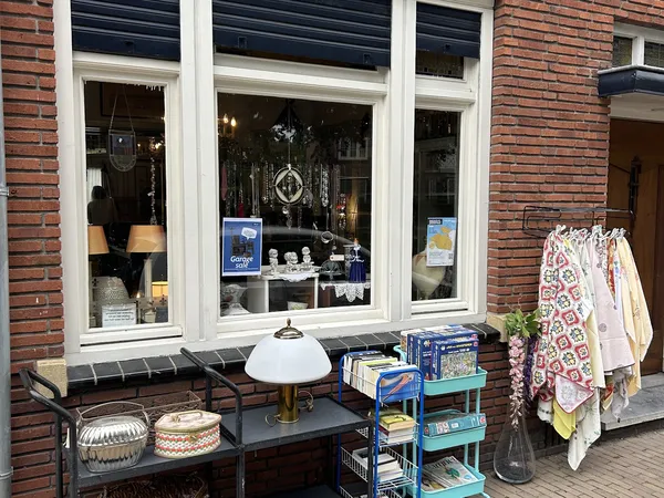 Winkelruit met witte frames van kringloopwinkel, uitgestalde kleding en huishoudelijke spullen op tafels buiten