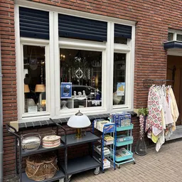 Winkelruit met witte frames van kringloopwinkel, uitgestalde kleding en huishoudelijke spullen op tafels buiten