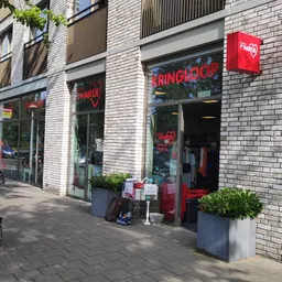 Gevel van kringloopwinkel met rode neonreclames en groene plantenbakken op straat