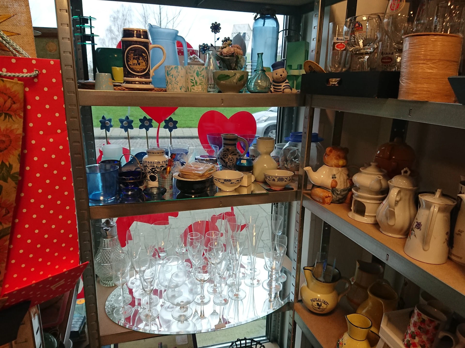 Glazen vitrinekast met servies, kannen, bekers en decoratieve items in een kringloopwinkel