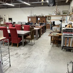 Ruim interieur van kringloopwinkel met diverse meubels: rood beklede stoelen, houten tafels, kasten en kleinere spullen op ve