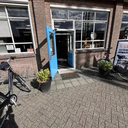Ingang van kringloopwinkel met blauwe deur, bakstenen gevel en ramen, fietsen en plantenbakken op het terras
