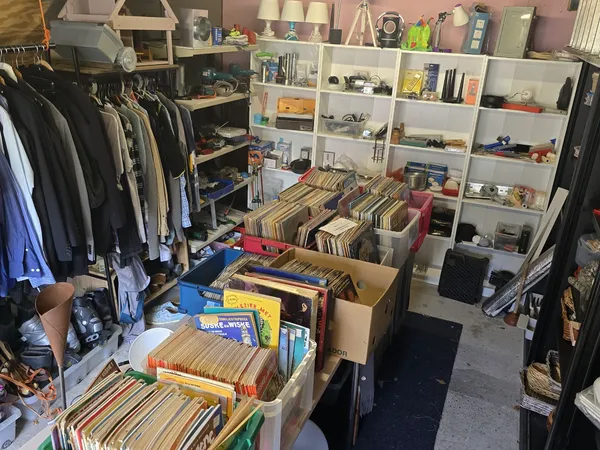 Winkelvloer met kledingrekken links, dozen met vinyl en media in het centrum, en witte stellingen met diverse spullen rechts