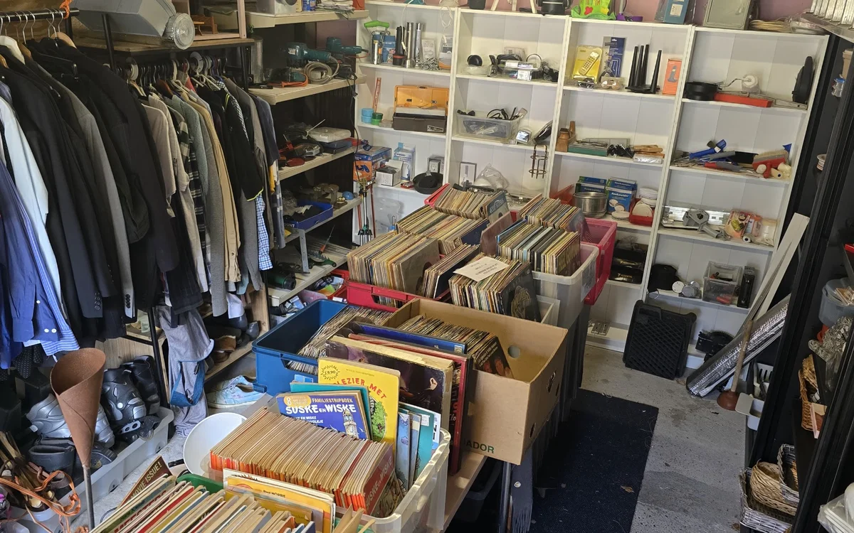 Winkelvloer met kledingrekken links, dozen met vinyl en media in het centrum, en witte stellingen met diverse spullen rechts