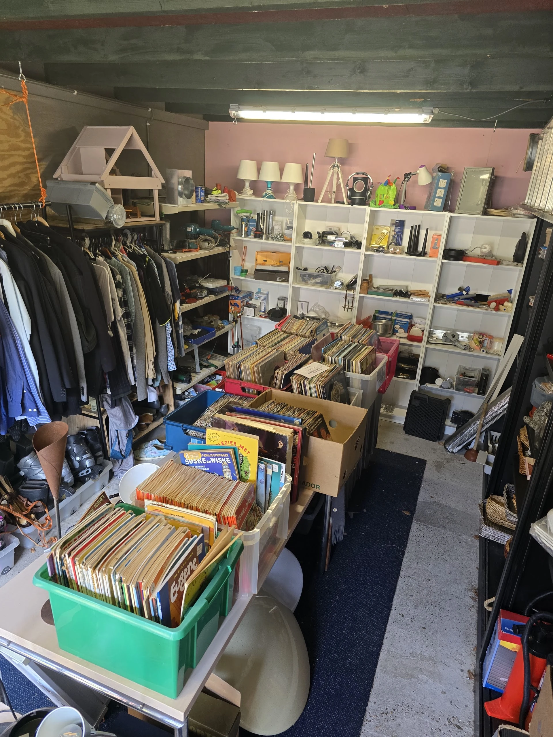 Winkelvloer met kledingrekken links, dozen met vinyl en media in het centrum, en witte stellingen met diverse spullen rechts