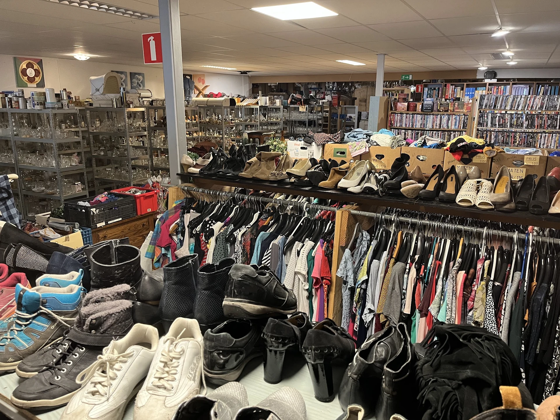 Rekken met tweedehands kleding, schoenen en tassen in een kringloopwinkel, met boeken op achtergrond