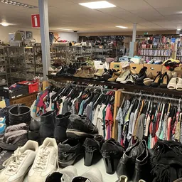 Rekken met tweedehands kleding, schoenen en tassen in een kringloopwinkel, met boeken op achtergrond