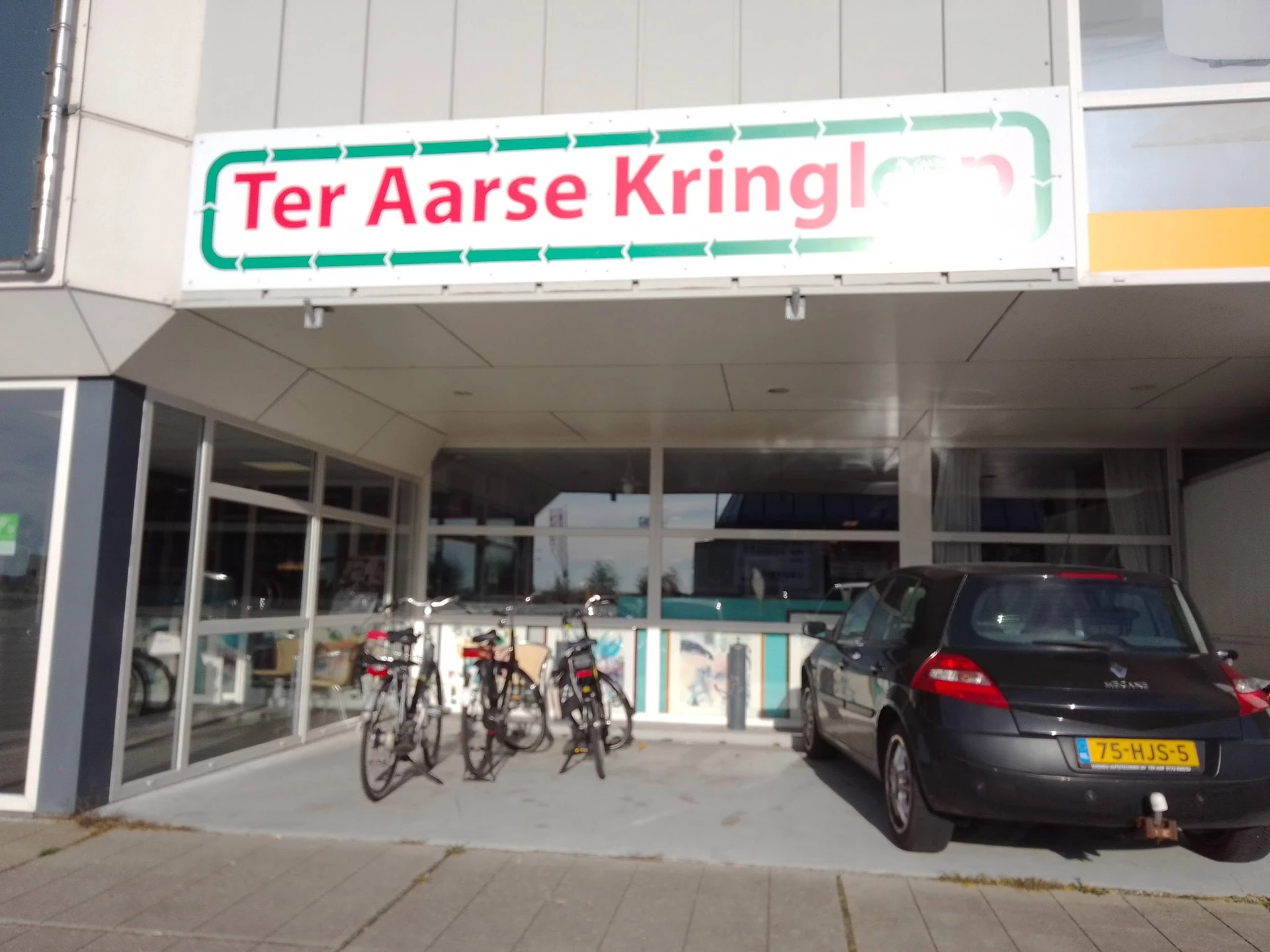 Gevel van Ter Aarse Kringloop met groene naam bord, glazen deuren en enkele fietsen ervoor
