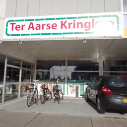 Gevel van Ter Aarse Kringloop met groene naam bord, glazen deuren en enkele fietsen ervoor