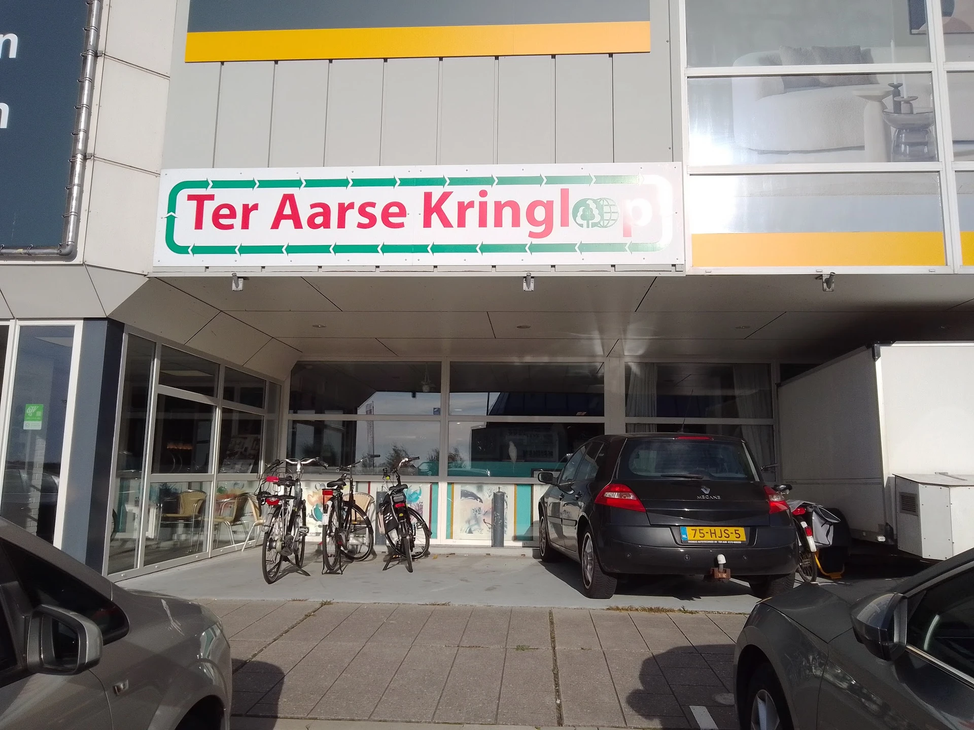 Winkelfront van Ter Aarse Kringloop met groene letters op witte achtergrond, ingang met glazen deuren, fietsen geparkeerd, au