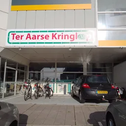 Winkelfront van Ter Aarse Kringloop met groene letters op witte achtergrond, ingang met glazen deuren, fietsen geparkeerd, au