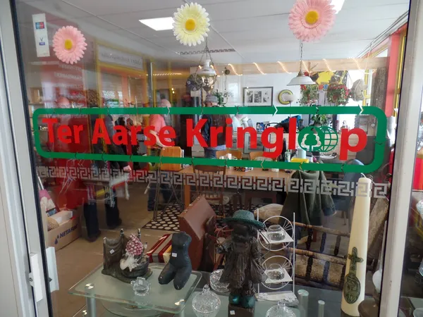 Winkelraam met groene winkelnaambord en decoratieve bloemen, uitzicht op winkelvloer met stellingen en spullen