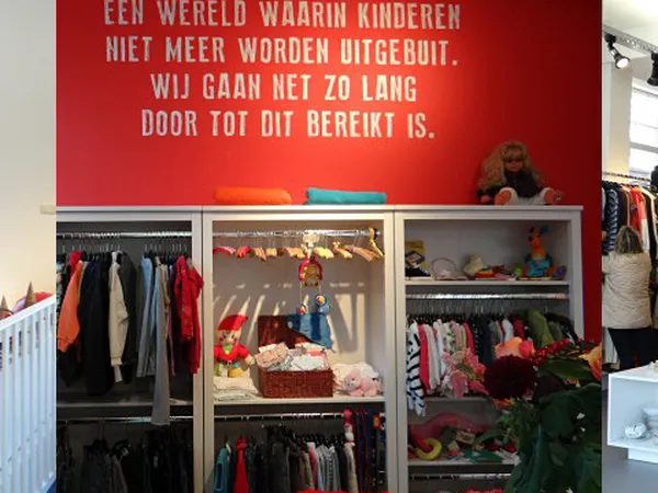 Kledingrekken en vitrines met kinderkleding en speelgoed in een kringloopwinkel, met een rood wandbord met Nederlandse tekst 