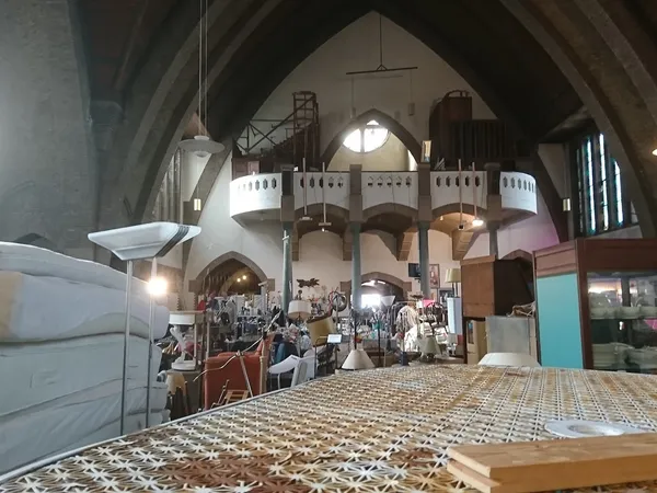Interieur van kringloopwinkel met hoge gewelfde plafonds, galerijen, diverse tweedehands spullen en kleding uitgestald op mee