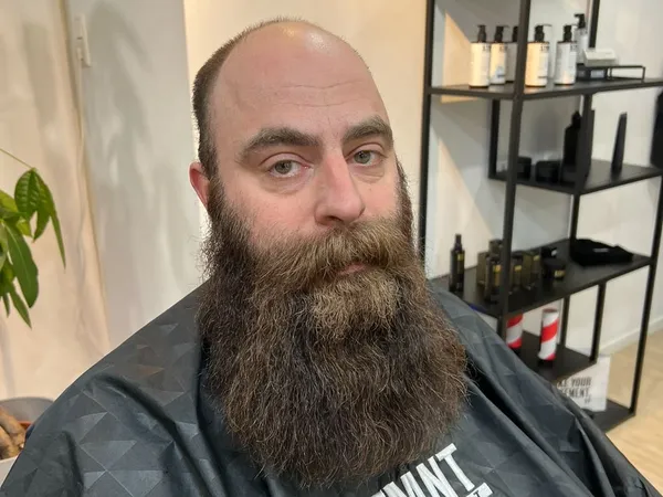 Man met lange baard in donker shirt in barbershop, stellingen met producten op achtergrond