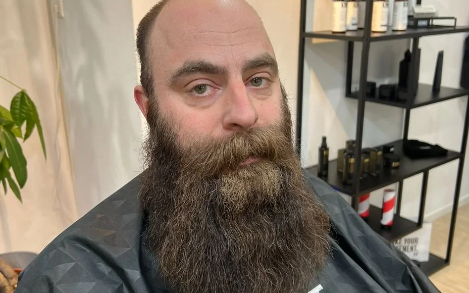 Man met lange baard in donker shirt in barbershop, stellingen met producten op achtergrond