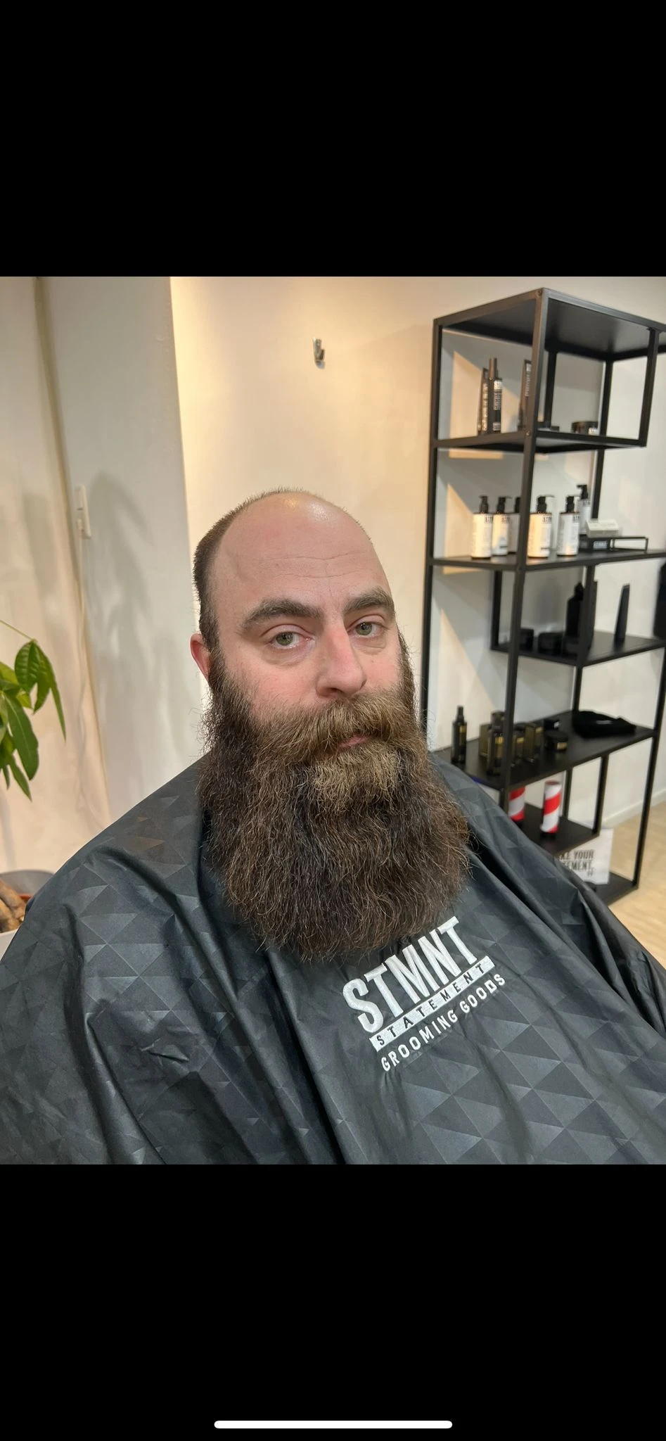 Man met lange baard in donker shirt in barbershop, stellingen met producten op achtergrond
