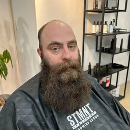 Man met lange baard in donker shirt in barbershop, stellingen met producten op achtergrond