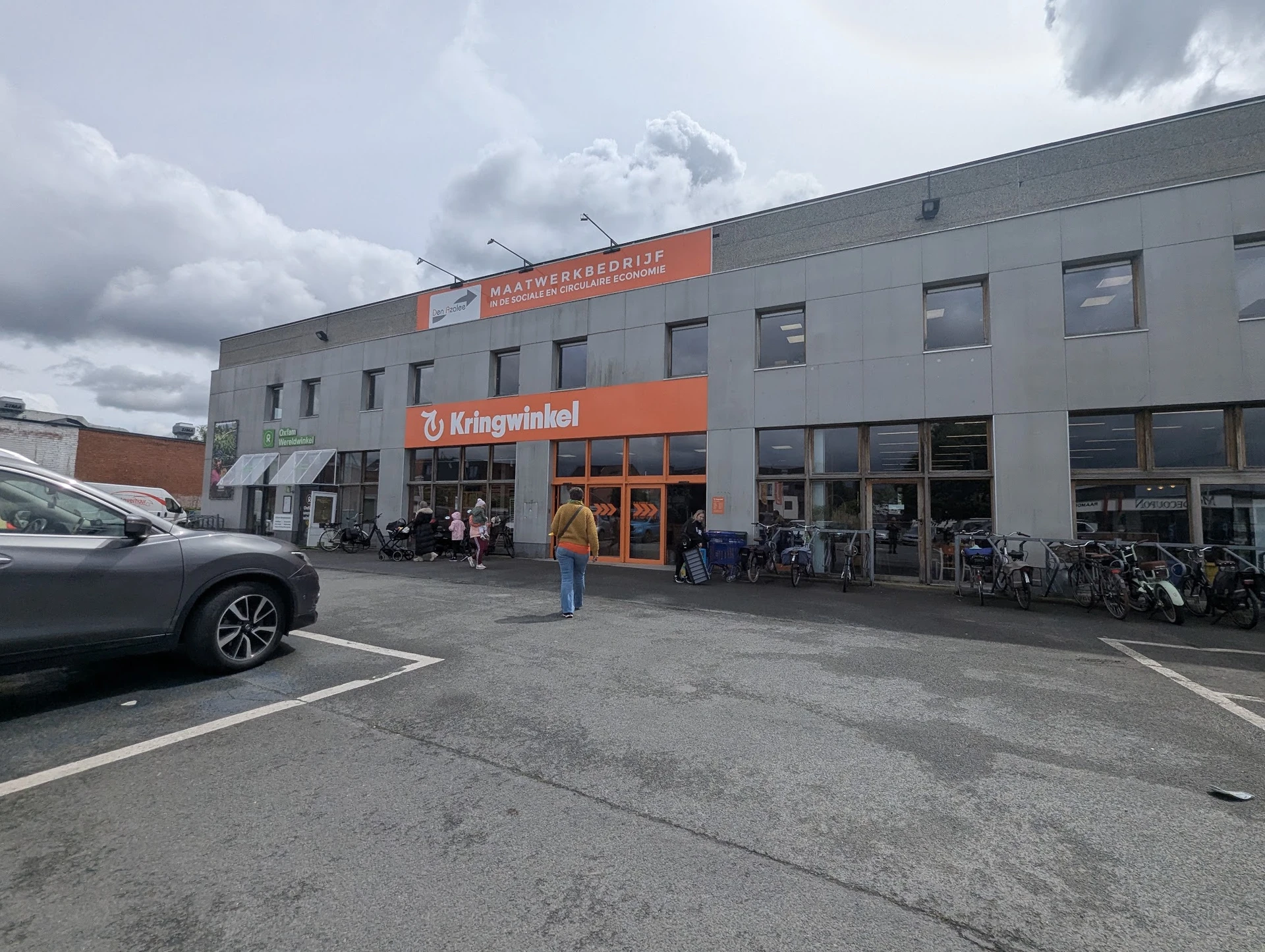 Gevel van kringloopwinkel Azalee met oranje uithangbord, ingang met glazen deuren en fietsen ernaast