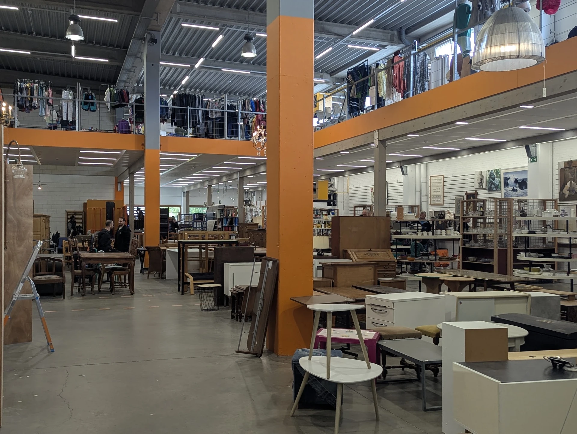 Ruim magazijn met diverse meubels, stoelen en huishoudelijke artikelen op display, ondersteund door oranje zuilen