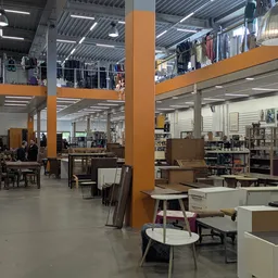 Ruim magazijn met diverse meubels, stoelen en huishoudelijke artikelen op display, ondersteund door oranje zuilen