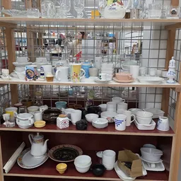 Glazen vitrine met diverse tweedehands serviesgoed, kopjes, kannen en potten