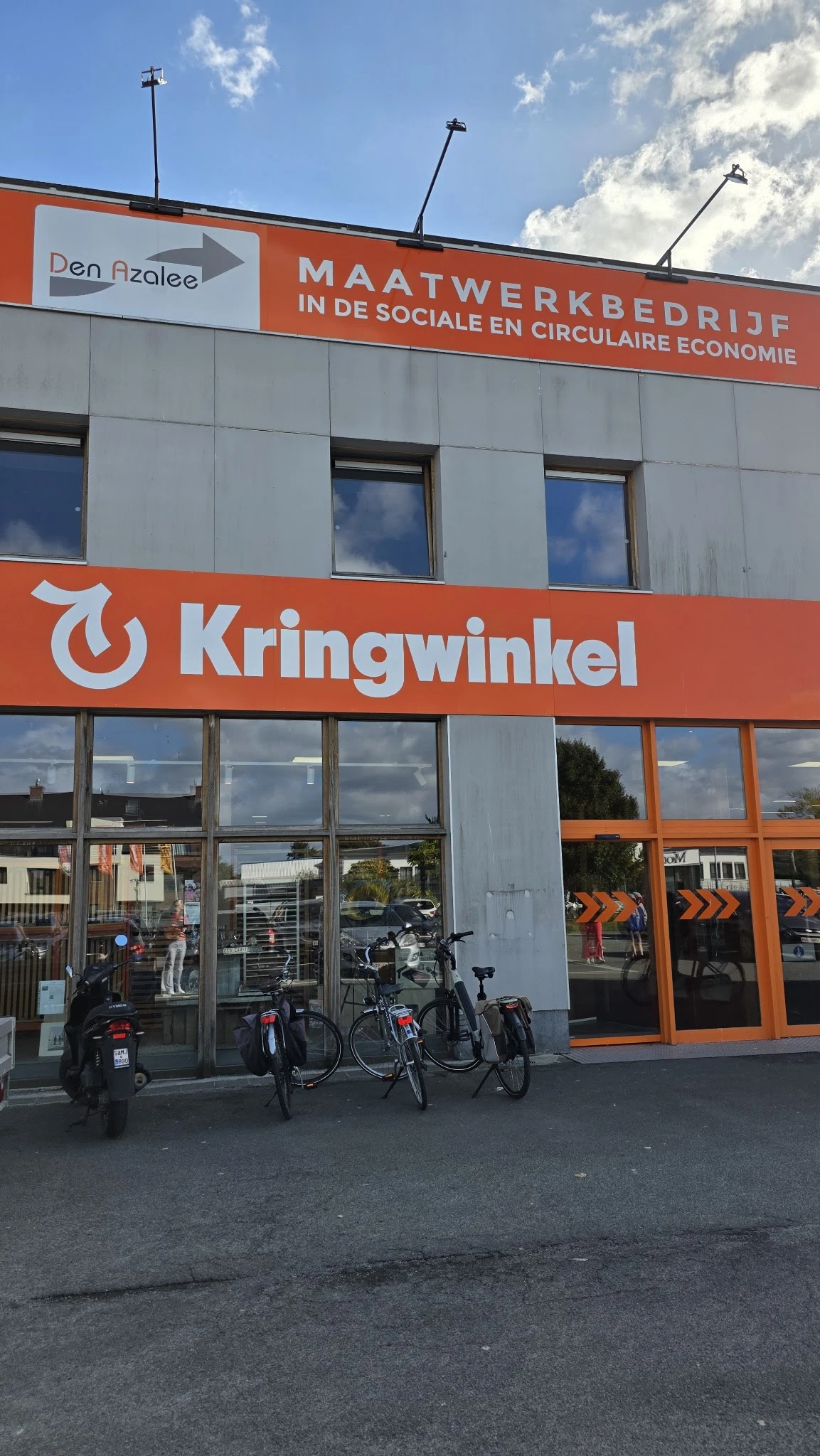 Winkelgevel met oranje Kringwinkel-logo en grote ramen, fietsen geparkeerd ervoor