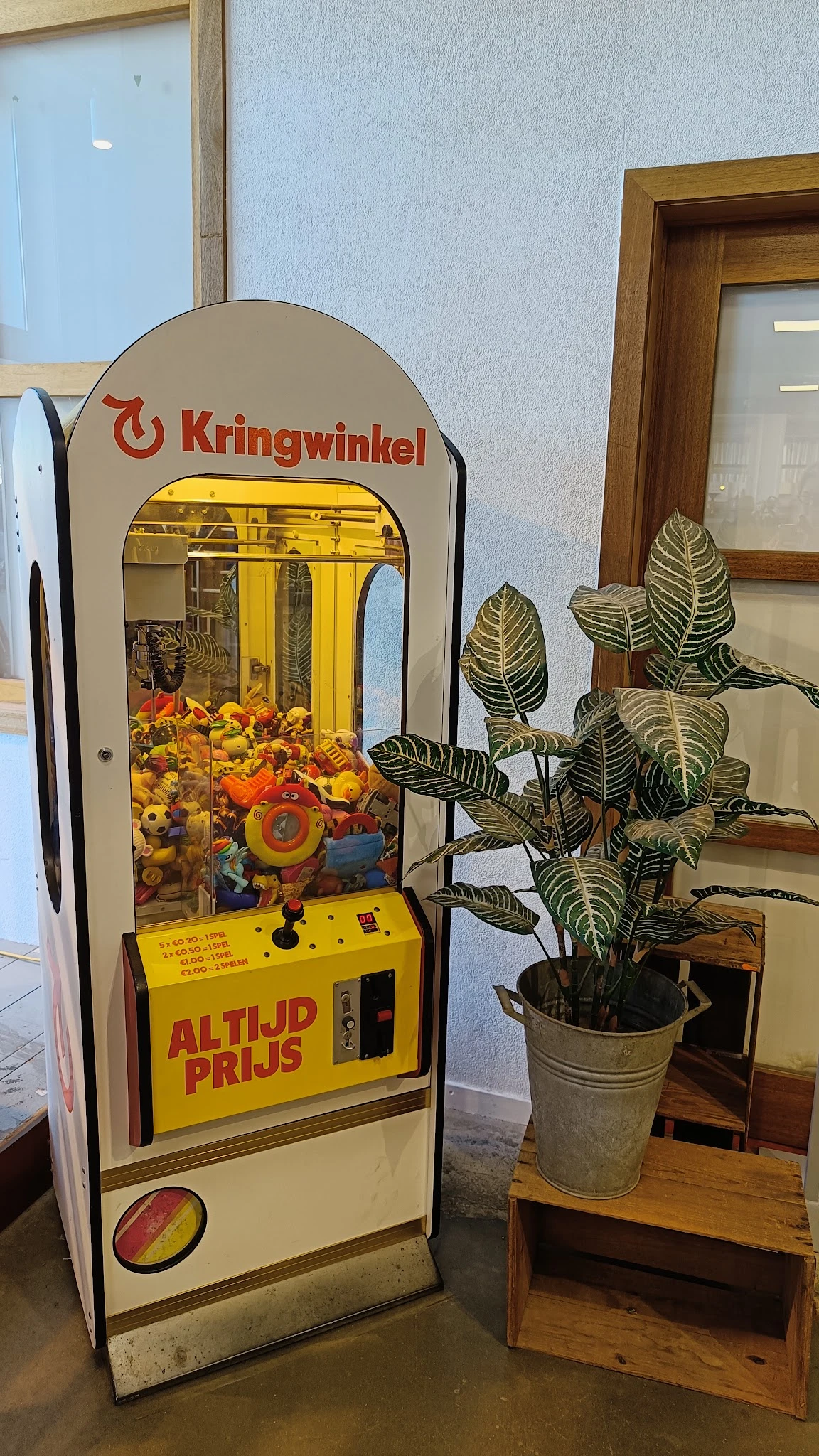 Grijparmenmachine met geel frame en speelgoedaanbod, potplant ernaast in winkelinterieur