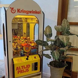 Grijparmenmachine met geel frame en speelgoedaanbod, potplant ernaast in winkelinterieur