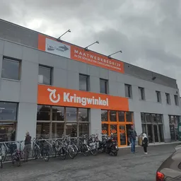 Gevel van kringloopwinkel met oranje Kringwinkel-logo, fietsenstalling en voetgangers op het plein